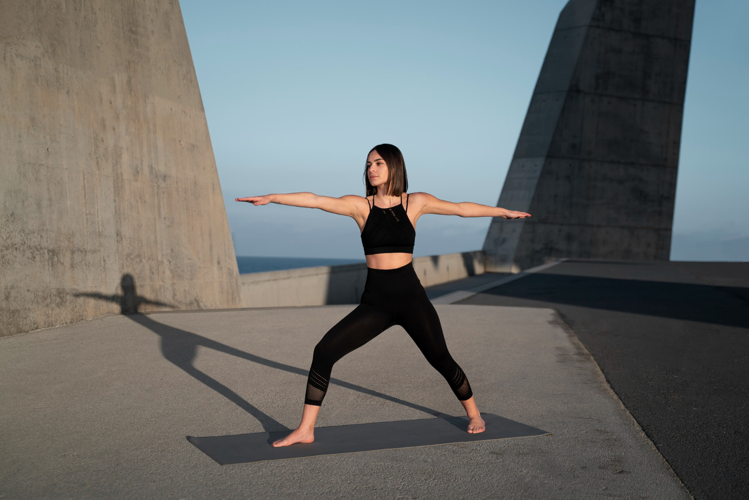 Yoga Mujer en la postura del guerrero II sobre una esterilla, en una azotea de hormigón junto al mar