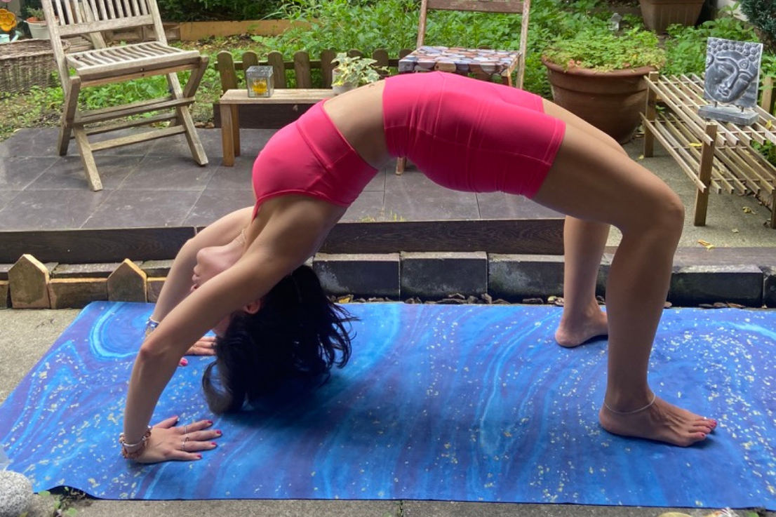 Yoga Mujer con ropa de yoga rosa en la postura de la rueda sobre una esterilla de viaje azul, en un jardín