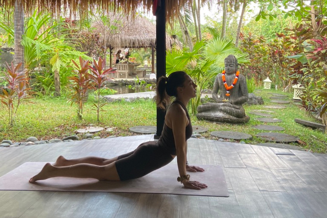 Yoga Mujer en la postura del perro boca arriba sobre una esterilla, en un pabellón abierto de Bali con jardín tropical y estatua de Buda al fondo