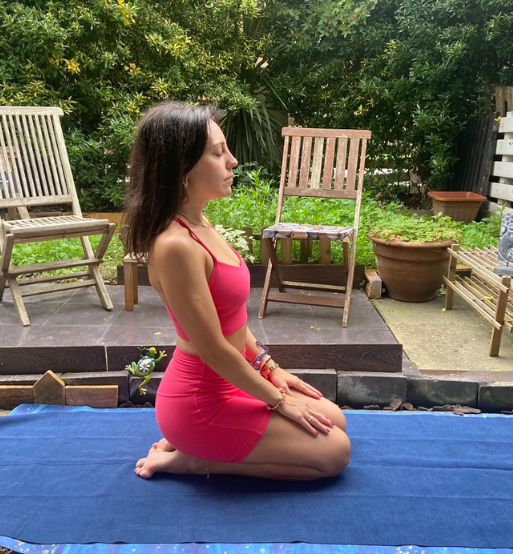 Laura sentada sobre los talones con los ojos cerrados, meditando en una esterilla azul en un jardín