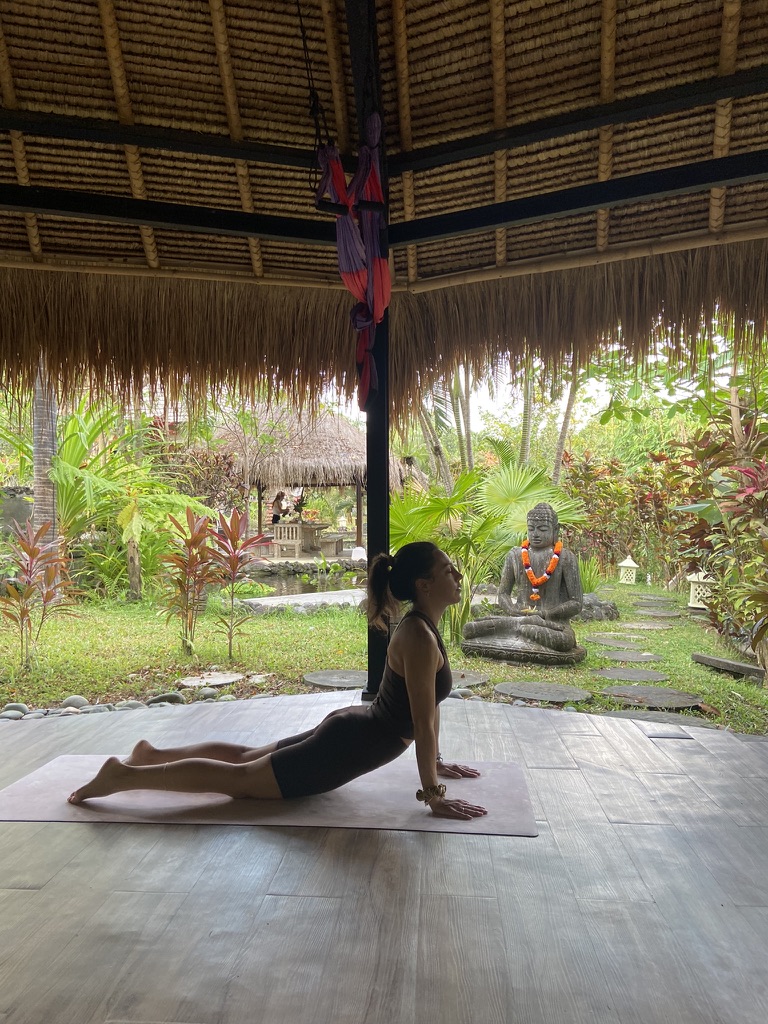 Mujer en postura de cobra sobre una esterilla en un pabellón abierto de Bali, con jardín tropical y estatua de Buda al fondo