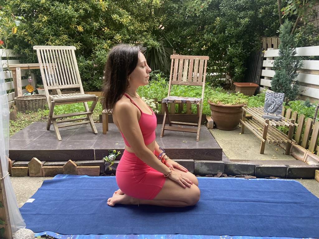 Mi reseña de la esterilla de yoga de viaje Mikkoa – Esterilla plegable para viajar Mujer en ropa de yoga rosa sentada sobre los talones meditando en una esterilla azul en un jardín