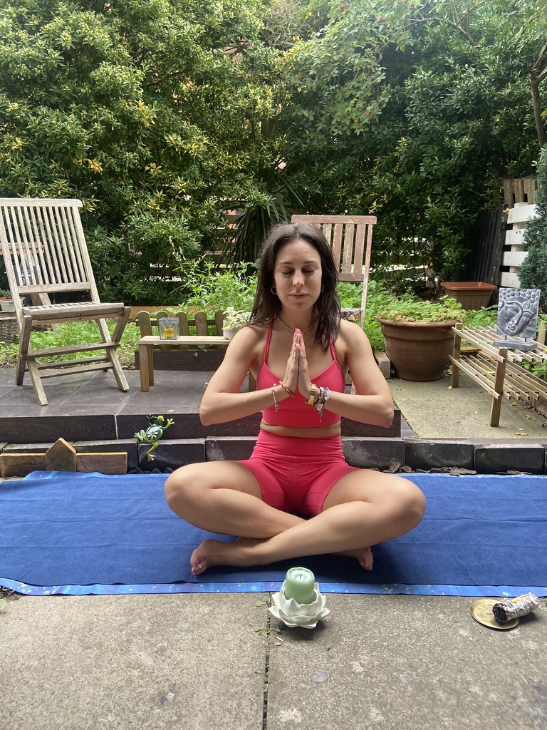 Mi reseña de la esterilla de yoga de viaje Mikkoa – Esterilla plegable para viajar Mujer en ropa de yoga rosa sentada con las piernas cruzadas y las manos juntas sobre una esterilla azul