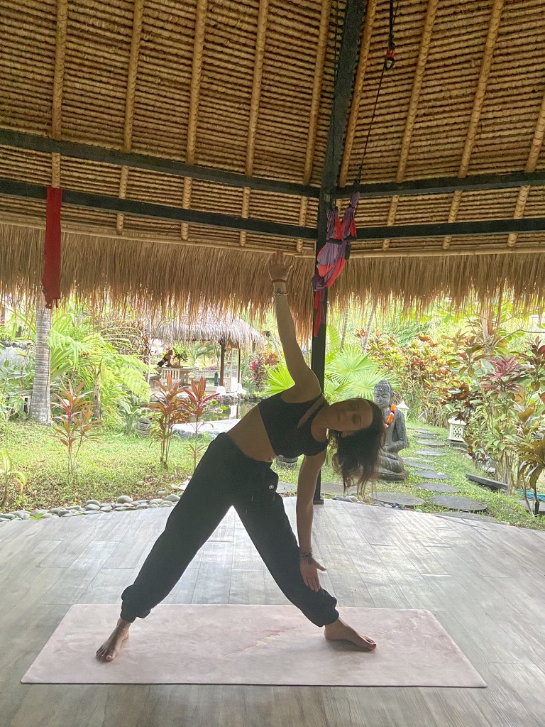 Mujer practicando yoga en la postura del triángulo en un pabellón de techo de paja rodeado de vegetación tropical en Bali