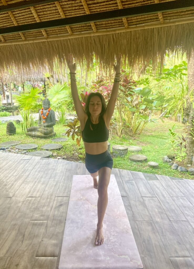 Laura practicando yoga sobre una esterilla en una terraza tropical de Bali, con jardín y estatua de Buda al fondo