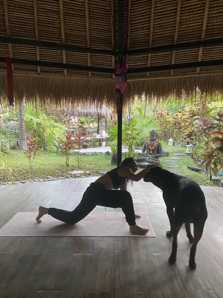 Mujer haciendo yoga en una esterilla acaricia a un perro negro en un pabellón abierto con jardín tropical y Buda al fondo