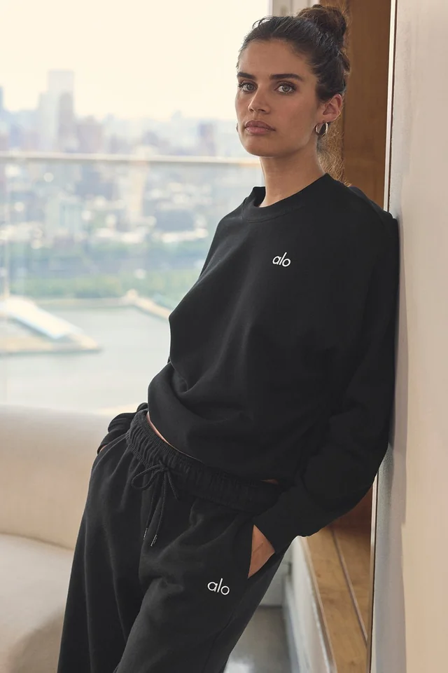 Modelo con sudadera y pantalón negros de Alo Yoga apoyada en una ventana con vistas a la ciudad