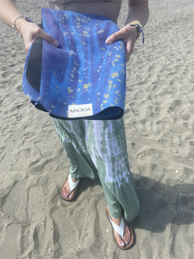 Mi reseña de la esterilla de yoga de viaje Mikkoa – Esterilla plegable para viajar Manos sosteniendo la esterilla de yoga de viaje Mikkoa plegada, con etiqueta visible, en la playa