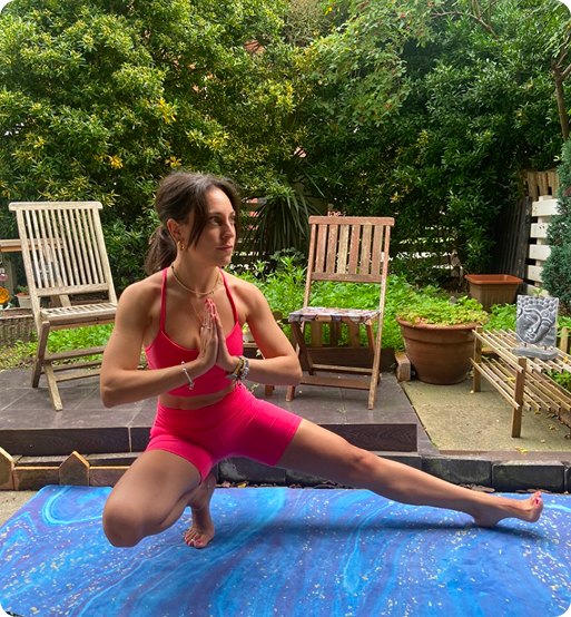 Laura en la postura de skandasana, con las manos juntas en anjali mudra sobre una esterilla azul en un jardín