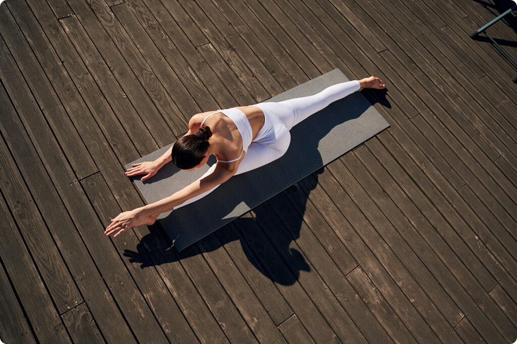 Mujer con conjunto de yoga blanco estirando el costado sobre una esterilla negra en una terraza de madera, vista desde arriba